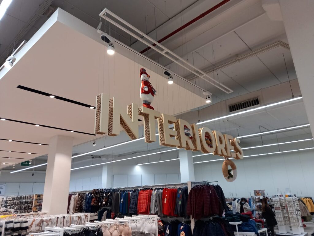 Letras caixa "INTERIORES" suspensas em placa estruturada — sinalização aérea de loja